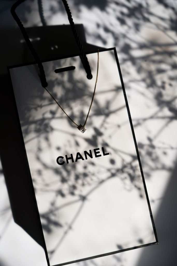 Stylish Chanel paper bag with a delicate necklace in artistic shadows on a sunny day.